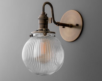May include: A vintage-style wall sconce with a clear, ribbed glass globe. The fixture has a brass finish, including the curved arm, bulb socket, and circular wall mount. An Edison-style bulb is visible inside the globe.