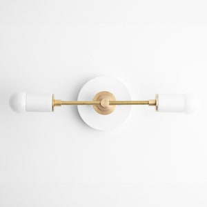 May include: A modern wall sconce with a white circular base and two white cylindrical light fixtures. The fixtures are connected by a brass rod and accented with brass details. The light bulbs are white and rounded.