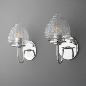 May include: Two wall sconces with clear, textured glass shades and chrome-finished metal hardware. The sconces have a classic design with a curved arm and a round backplate. The light bulbs are visible inside the glass shades.