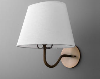 May include: A wall-mounted lamp with a white lampshade and a bronze-colored metal arm and base. The lampshade is a classic tapered shape. The lamp is designed to be mounted on a wall. The lamp's dimensions are not visible.