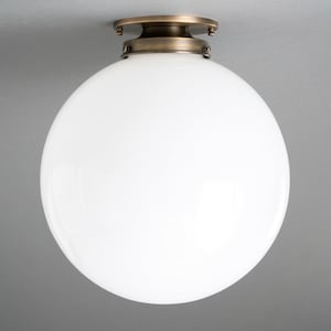 May include: A white globe ceiling light fixture with a brass-colored base. The round, opaque glass shade is illuminated, and the fixture is mounted on a gray ceiling. This modern light fixture provides ambient lighting.
