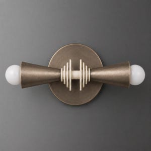 May include: A wall sconce featuring a circular bronze backplate and two horizontal, cone-shaped arms extending outward. The center displays a series of stacked, thin, light-colored circular discs, and each end is capped with a round white light bulb.