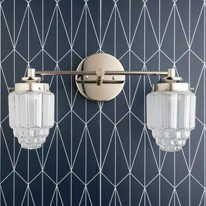 May include: A two-light vanity fixture with a polished nickel finish. The fixture features two frosted glass shades with vertical ribbed detailing. The fixture is mounted on a circular backplate and a horizontal bar. The background is a navy blue wall with a white geometric pattern.