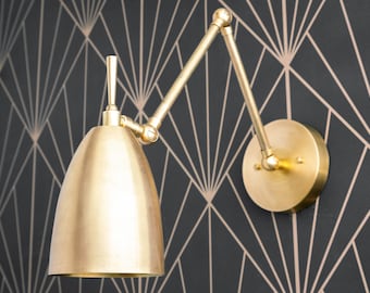 May include: A brass wall lamp featuring a dome-shaped shade and an adjustable articulated arm with multiple joints. The fixture includes a circular wall mounting plate and a vertical handle on top of the shade for positioning the light.