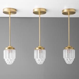 May include: Three identical pendant lights featuring a brushed brass finish on the circular ceiling canopy and thin vertical rod. Each fixture includes a clear, faceted glass shade with a tiered, geometric design that tapers toward the bottom.