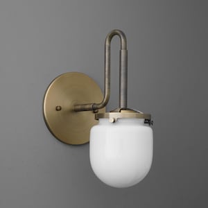 May include: A wall-mounted light fixture with a brass-colored circular base and curved arm. The fixture holds a white, rounded glass shade. The metal components have an aged appearance, suggesting a vintage or industrial design. The fixture is mounted on a gray wall.
