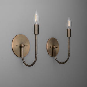 May include: Two antique brass wall sconces with clear, elongated light bulbs. Each sconce has a circular backplate, a curved arm, and a vertical bulb holder. The sconces are mounted on a gray wall.