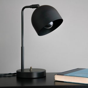 May include: A black metal desk lamp with a round shade and a black cord. The lamp is on a black surface next to a blue book.
