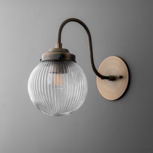 May include: A vintage-style wall sconce with a clear, ribbed glass globe and a bronze-colored metal frame. The fixture has a curved arm and a circular wall mount, showcasing an Edison-style bulb.