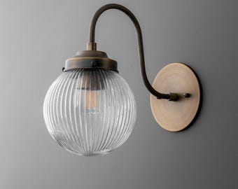 May include: A vintage-style wall sconce with a clear, ribbed glass globe and a bronze-colored metal frame. The fixture has a curved arm and a circular wall mount, showcasing an Edison-style bulb.