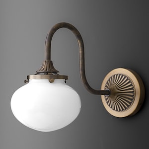 May include: A vintage-style wall sconce with a white glass shade and a curved, bronze-colored arm. The sconce has a decorative, circular wall mount with a sunburst design. The overall design is reminiscent of antique lighting fixtures.