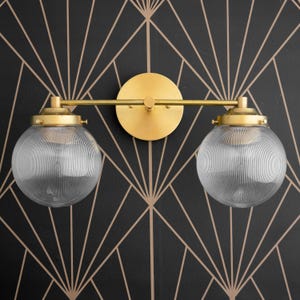 May include: A gold-toned wall sconce with two clear glass globe shades. The sconce has a simple, modern design and is perfect for adding a touch of elegance to any room.