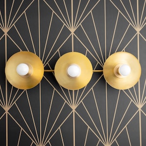May include: Three gold-toned light fixtures with white light bulbs mounted on a black wall with a geometric pattern. The fixtures are round and have a slightly curved shape.