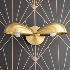 May include: A gold-toned wall sconce with two light bulbs. The sconce has a unique design with two curved shades that resemble seashells.