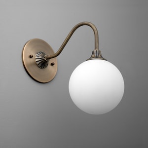 May include: A wall-mounted light fixture with a brass finish. It features a round backplate, a curved arm, and a white globe-shaped shade. The fixture is designed for indoor use and adds a vintage touch to any space.