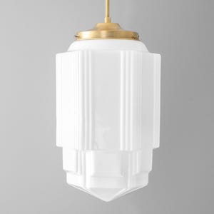 May include: A white, Art Deco-style pendant light fixture with a brass-colored top. The glass shade has vertical ribbed detailing and a geometric, tiered shape. The light hangs from a thin, gold-colored rod.