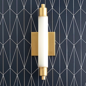 May include: A gold wall sconce with a white frosted glass cylinder light fixture. The sconce is mounted on a dark blue geometric patterned wall.