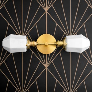 May include: A gold wall sconce with two white frosted glass shades. The sconce has a geometric design and is mounted on a black background.