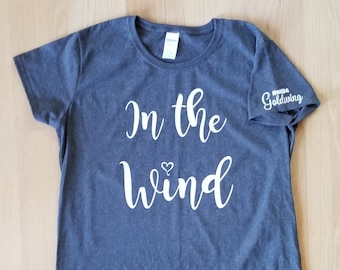 May include: A dark gray t-shirt with the text "In the wind" in white cursive lettering and a small heart symbol. The shirt has a small white logo on the sleeve that says "Honda Goldwing."