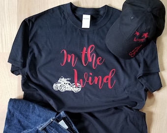May include: Black t-shirt with the text "In the wind" in red lettering and a graphic of a motorcycle. A black baseball cap with the text "In the wind" and "Spyder" in white lettering.