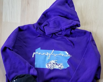 May include: Purple hoodie with a blue outline of the state of Pennsylvania and the word "Pennsylvania" written in white script. A black baseball cap with a white graphic of a motorcycle is on the hoodie.