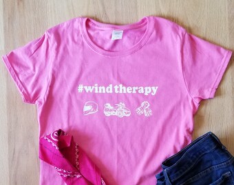 May include: Pink t-shirt with the text "#windtherapy" in white. The shirt also features a white helmet, a white ATV, and a pair of white gloves.