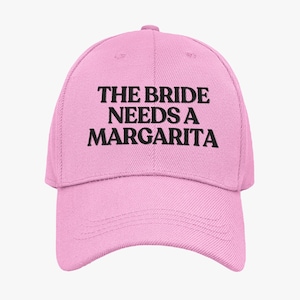 May include: A pale pink baseball cap featuring the black text "THE BRIDE NEEDS A MARGARITA". The cap has a curved peak and an adjustable strap. The hat is made of a soft, durable material, suitable for outdoor wear.