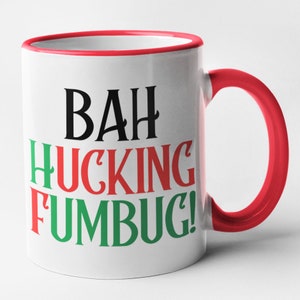May include: White ceramic mug with a red handle and rim. The mug features the words "BAH HUCKING FUMBUG!" in black, red, and green lettering. The mug is designed for hot beverages.