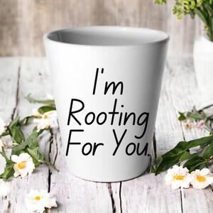 May include: A white ceramic plant pot with the black text "I'm Rooting For You". The pot is cylindrical and stands on a distressed white wooden surface, with greenery and white flowers around it.