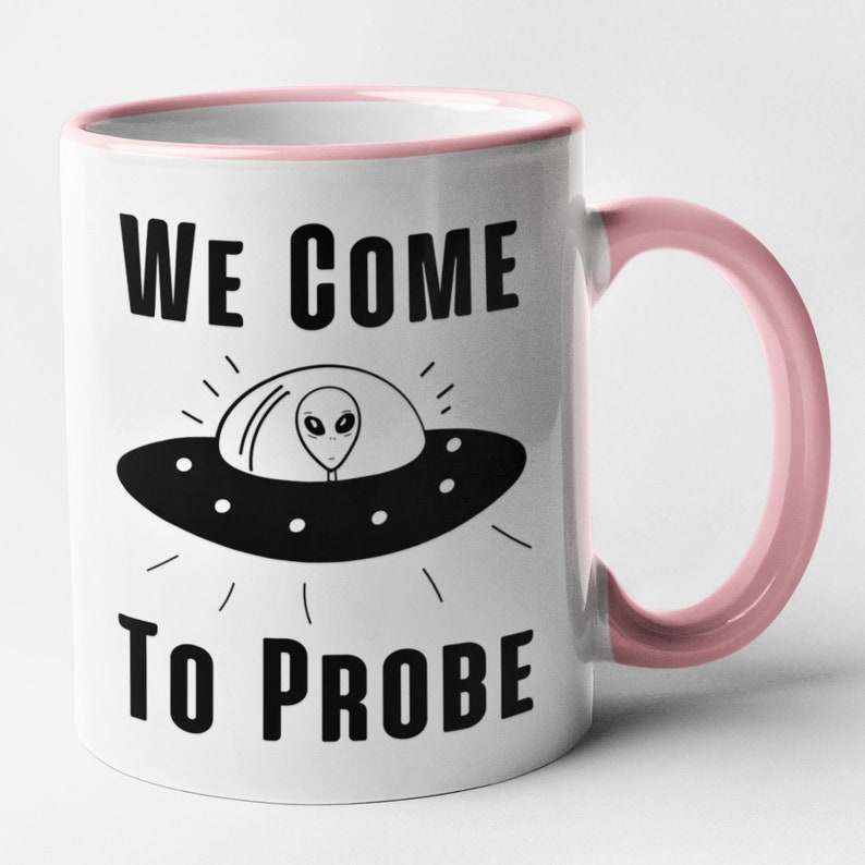 Op de afbeelding: Witte keramische koffiemok met een roze rand en handvat. De mok heeft een zwart-witte illustratie van een UFO met een alien erin en de tekst "WE COME TO PROBE".