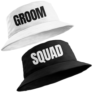 May include: Two bucket hats, one white and one black. The white hat features the word "GROOM" in large, bold, black letters. The black hat displays the word "SQUAD" in large, bold, white letters. Both hats are made of a soft fabric.