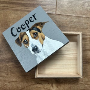 May include: A small wooden box with a hinged lid, featuring a painted portrait of a dog. The dog is depicted with white, brown, and black fur, and the name "Cooper" is written above. The box is grey, with a natural wood interior.