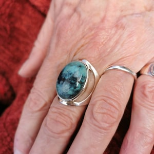 May include: A silver ring featuring a large oval turquoise stone set in a bezel setting. The ring is worn on a person's finger.