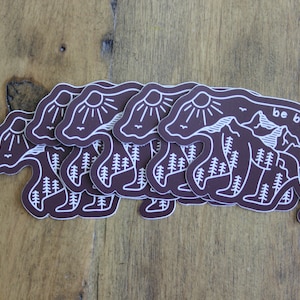 May include: A stack of brown bear stickers with a white outline. The bear is standing on a mountain with a sun and birds in the sky. The text "be brave" is on the right side of the sticker.