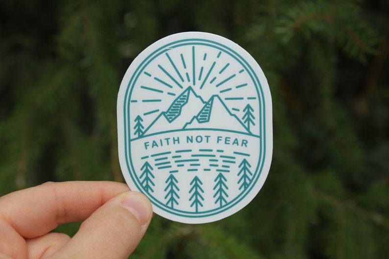 May include: A white sticker with a teal outline and design. The design features mountains, trees, and sun rays, with the words "FAITH NOT FEAR" in a bold font. The sticker is held by a hand.
