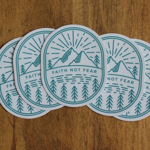 May include: Oval stickers with a teal and white mountain and forest design. The stickers have the text "FAITH NOT FEAR" in a sans-serif font. The stickers are arranged on a wooden surface.