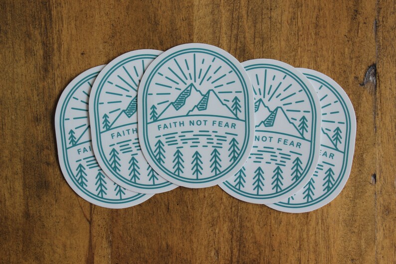 May include: Oval stickers with a teal and white mountain and forest design. The stickers have the text "FAITH NOT FEAR" in a sans-serif font. The stickers are arranged on a wooden surface.