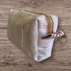 Op de afbeelding: Een rechthoekige, beige toilettas met een bruine rits en een gestreepte handgreep. De tas is gemaakt van een textuurstof met verticale bruine strepen. De rits is lichtbruin.