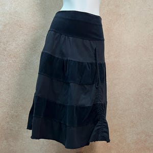 May include: A black knee-length skirt featuring a wide waistband and alternating horizontal panels of smooth and textured fabric. The skirt includes a vertical drawstring detail on the right side, creating a gathered effect along the hemline.