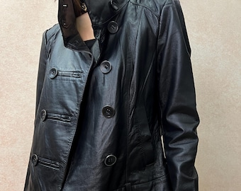 May include: A black leather jacket featuring a double-breasted front closure with large round buttons, a high stand-up collar, long sleeves, and a cropped hemline. The jacket is worn over blue acid-wash denim jeans with visible silver hardware.