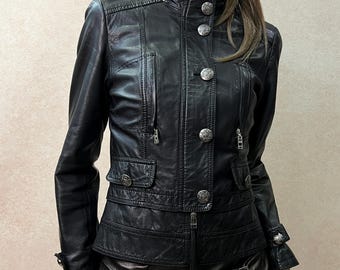 May include: A black leather jacket featuring a high collar, silver-toned embossed buttons, and two front flap pockets. The jacket is paired with a matching black leather mini skirt that has an asymmetrical wrap-style front with silver metal buckles.