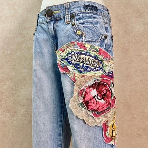 Könnte beinhalten: Hellblaue Jeans aus Denim mit einer großen, mehrlagigen Stoffapplikation auf dem linken Oberschenkel. Die Applikation besteht aus rosa, roten und cremefarbenen Spitzen- und Stoffresten mit dem gestickten Text JEANS und einem roten STOP-Schild.