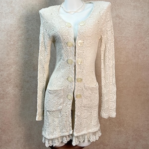 May include: A long-sleeved, cream-colored cardigan featuring an intricate open-knit crochet pattern. The garment has a round neckline, four large circular buttons down the front, two front patch pockets, and a delicate lace trim along the hem.