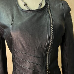 May include: A black leather jacket featuring a round neckline, long sleeves, and an asymmetrical silver-tone metal zipper closure that runs diagonally across the front. The jacket includes horizontal stitched panel detailing on the torso.