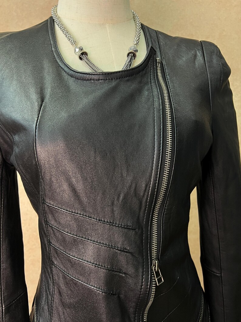 May include: A black leather jacket featuring a round neckline, long sleeves, and an asymmetrical silver-tone metal zipper closure that runs diagonally across the front. The jacket includes horizontal stitched panel detailing on the torso.