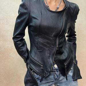 May include: A black leather jacket featuring an asymmetrical front zipper closure, long sleeves, and two zippered pockets at the waist. The jacket has a round neckline and structured shoulders, paired with blue denim jeans with a visible metal button.