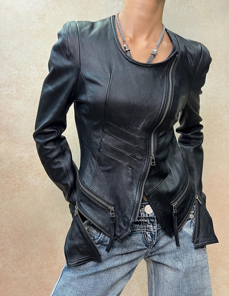 May include: A black leather jacket featuring an asymmetrical front zipper closure, long sleeves, and two zippered pockets at the waist. The jacket has a round neckline and structured shoulders, paired with blue denim jeans with a visible metal button.