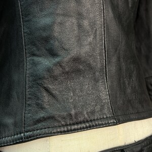 May include: A black leather jacket featuring a smooth, textured surface with visible vertical seam detailing. The garment is constructed with durable stitching along the hemline and side panels, showcasing a matte finish and natural grain.