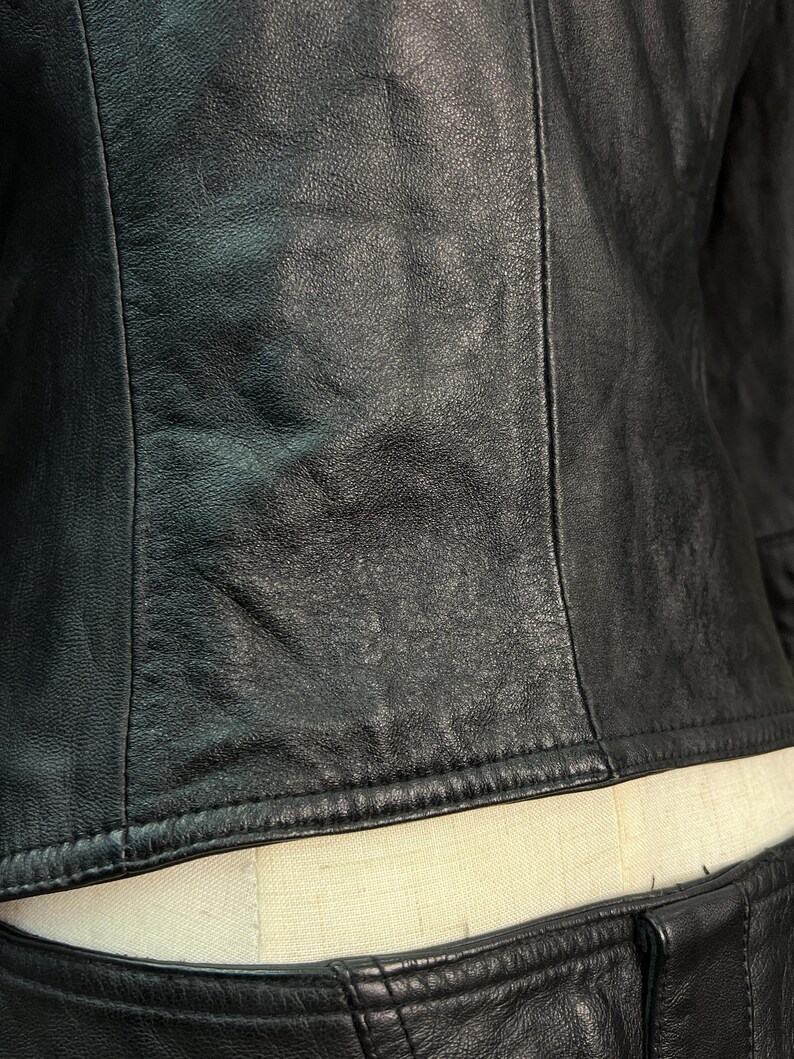 May include: A black leather jacket featuring a smooth, textured surface with visible vertical seam detailing. The garment is constructed with durable stitching along the hemline and side panels, showcasing a matte finish and natural grain.