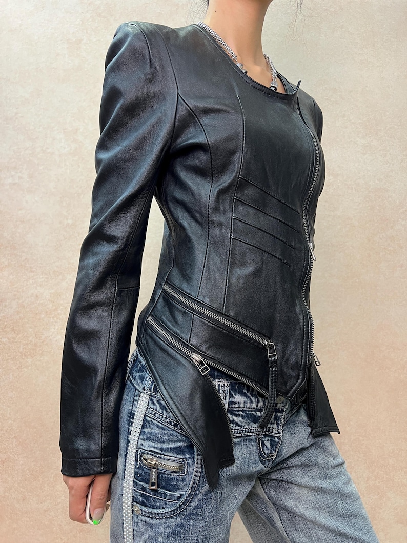 May include: A black leather jacket featuring a round neckline, long sleeves, and a full-length front zipper. The jacket includes horizontal stitched detailing on the chest and two diagonal zippered pockets at the waist with an asymmetrical hemline.